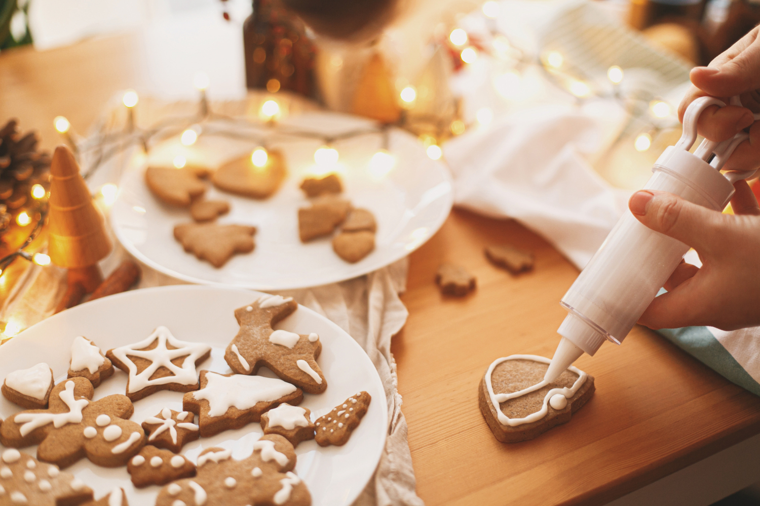 Blogue activités pour se mettre dans l’esprit des fêtes (2) Personne décorant des biscuits de Noël en pain d'épices avec du glaçage blanc, entourée de guirlandes lumineuses et de décorations festives. L'ambiance est chaleureuse et met en avant les préparatifs des fêtes.