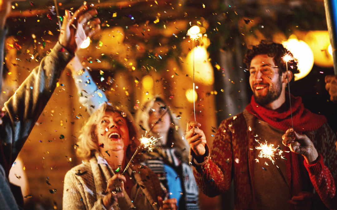 Un groupe de personnes souriantes célèbre sous une pluie de confettis et d’étincelles dans une ambiance festive et chaleureuse. Ils tiennent des cierges magiques qui illuminent la scène, tandis que l’arrière-plan dévoile des lumières tamisées et une décoration accueillante. L’énergie joyeuse et conviviale de cette image évoque une fête de fin d’année ou une célébration du Nouvel An en plein air.