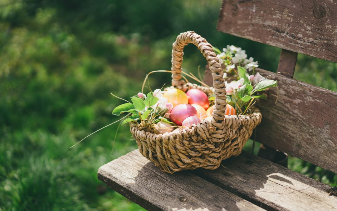 Un panier en osier rempli d’œufs de Pâques colorés et de fleurs printanières, posé sur un banc en bois dans un jardin, illustrant des idées originales pour célébrer Pâques en plein air.