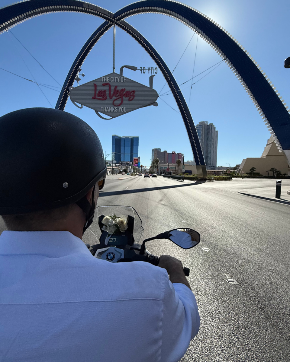 Vue depuis l’arrière d’un scooter décoré de fleurs blanches, conduit sous l’arche emblématique de Las Vegas. La photo illustre un couple fraîchement marié profitant de son mariage à Vegas, symbole d’aventure et de liberté.