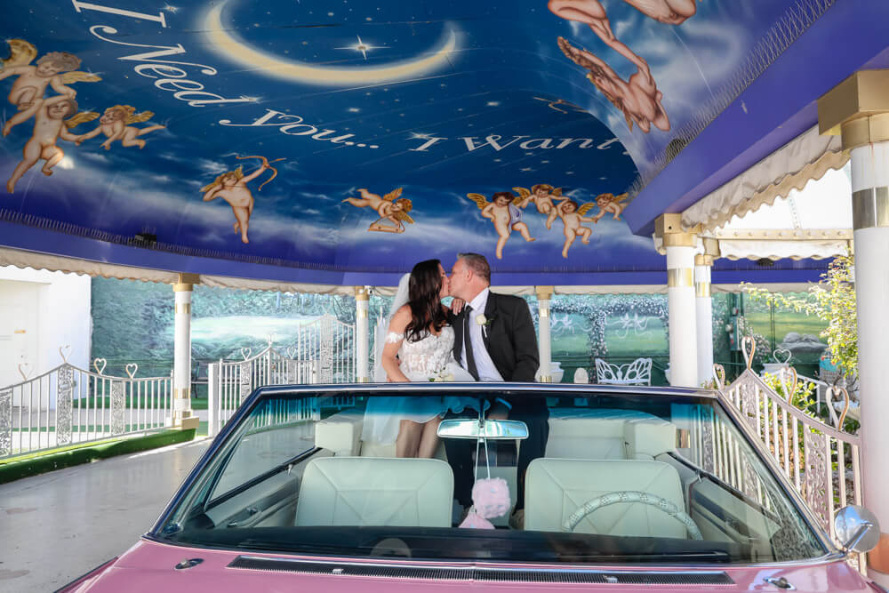 Un couple s’embrasse dans une Cadillac rose lors de leur mariage à Vegas, sous le célèbre auvent peint de la Little White Chapel orné de chérubins et de la phrase “I Need You… I Want You.” L’image capture toute la magie et la fantaisie d’un mariage à Las Vegas.