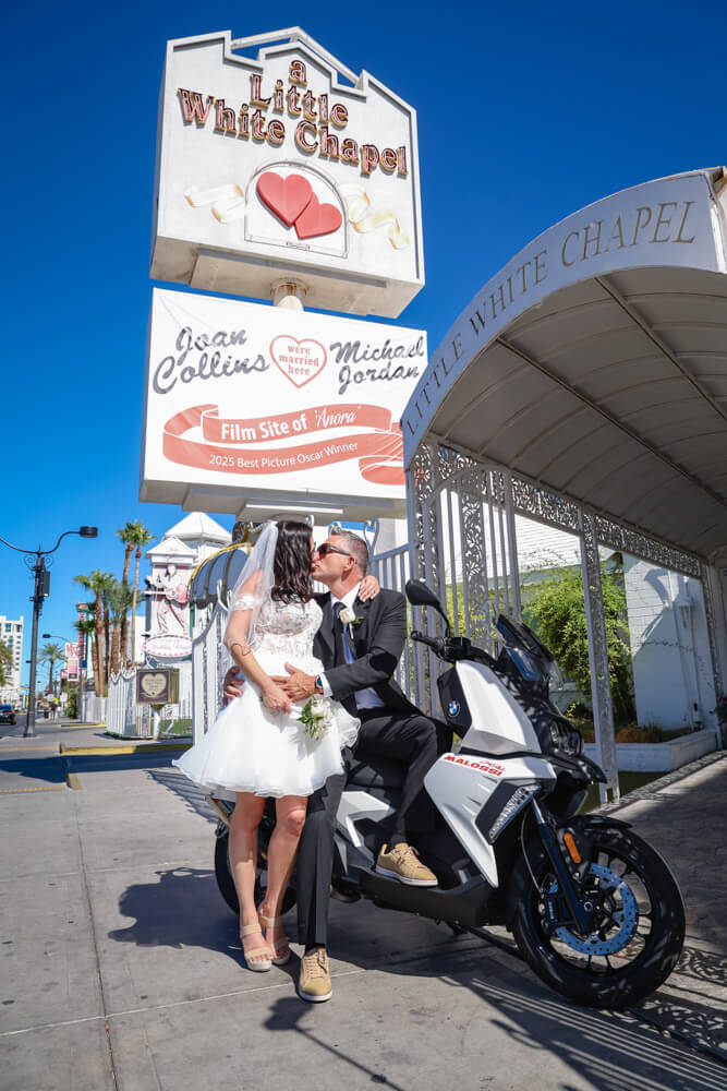 Un couple pose devant la célèbre Little White Chapel pour leur mariage à Vegas. La mariée en robe blanche et le marié en complet noir s’embrassent près d’un scooter, sous le ciel bleu de Las Vegas.