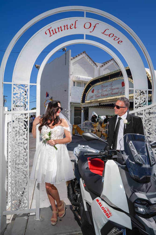 Un couple fraîchement marié pose sous l’arche « Tunnel of Love » de la Little White Chapel, symbole emblématique du mariage à Vegas. La mariée en robe blanche et le marié en complet noir se tiennent près d’un scooter, rayonnant de bonheur après leur cérémonie romantique et spontanée.